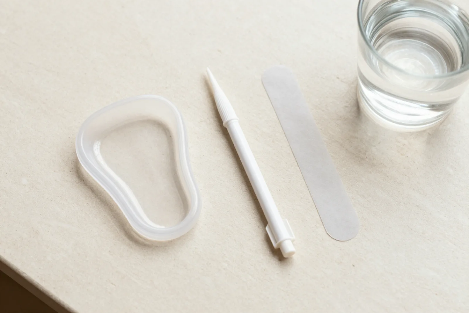Tandblekningsprodukter uppradade uppifrån på en ljus bänkskiva: skena, penna, remsor och ett glas vatten.